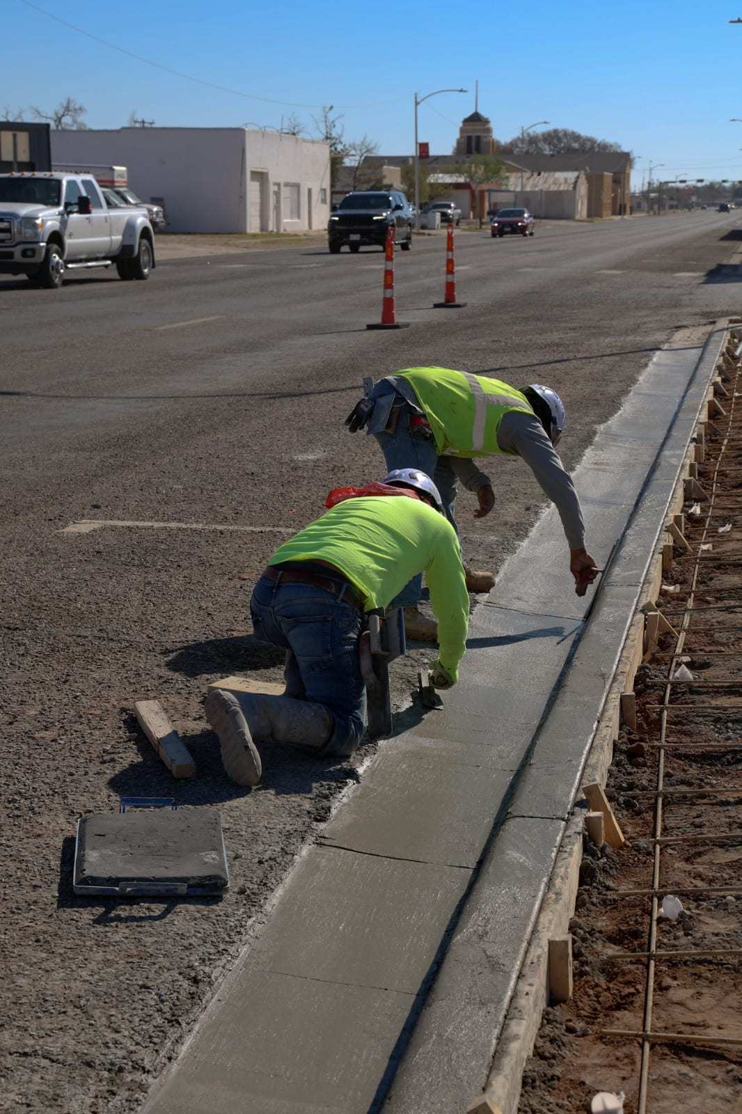 Two crew members in safety vests kneeling to finish a freshly placed concrete curb and gutter beside a road