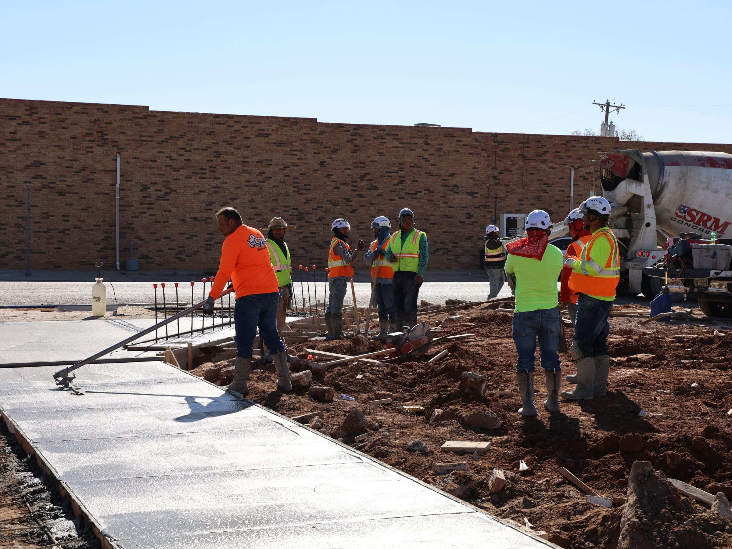Crew screeding a fresh concrete flatwork pour beside a brick building, with a ready-mix truck in the background