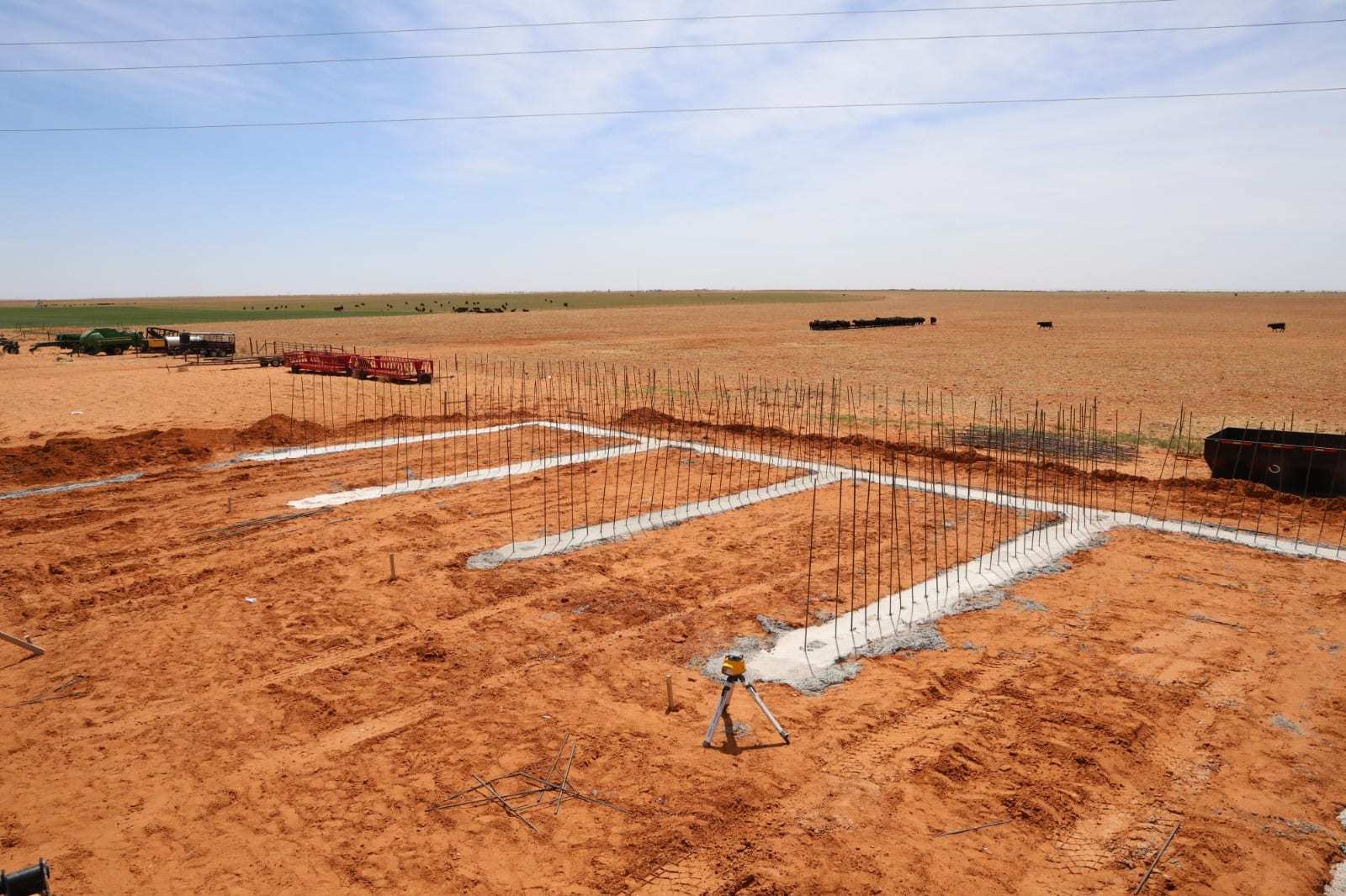 Concrete footings with rows of vertical rebar dowels laid out for stem walls, with a laser level on a tripod in the foreground
